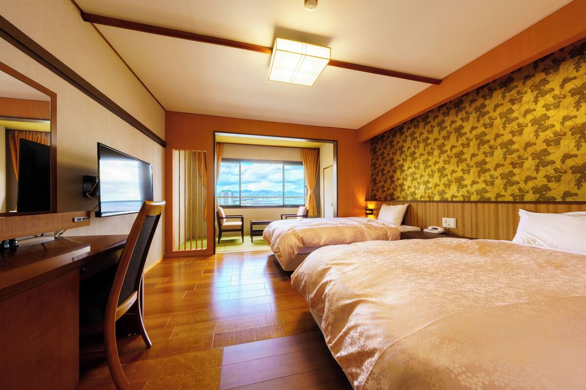 A hotel room with two beds, a desk with a chair, a television, and a large window with curtains.