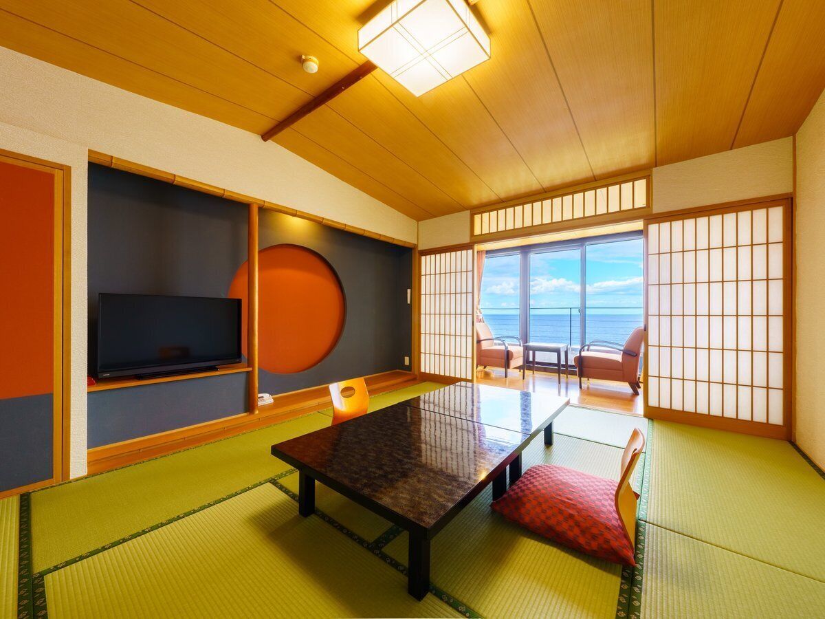 A traditional Japanese room with tatami flooring, a low table, a television, and a view of the sea.