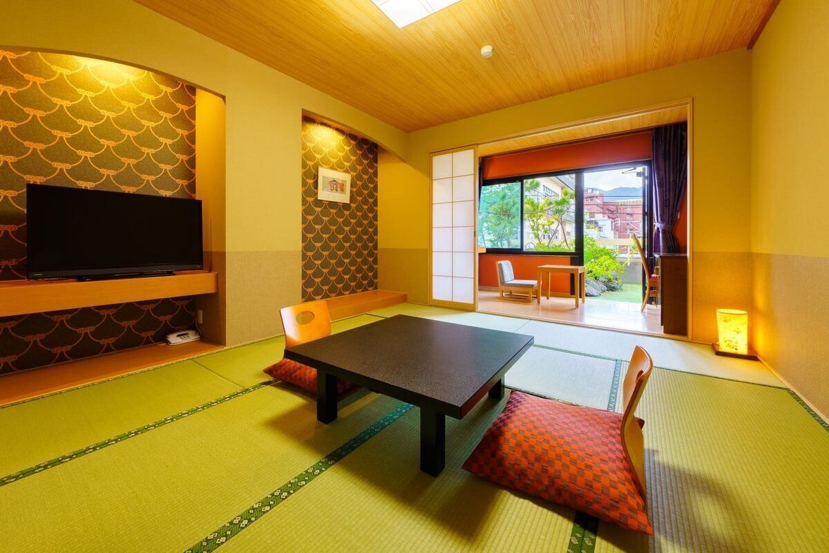 A tatami mat room with a low table, cushions, a television, and sliding doors leading to an outdoor area.