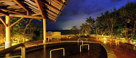 A pool area with a wooden pergola and railings, surrounded by trees and a view of the ocean at night.