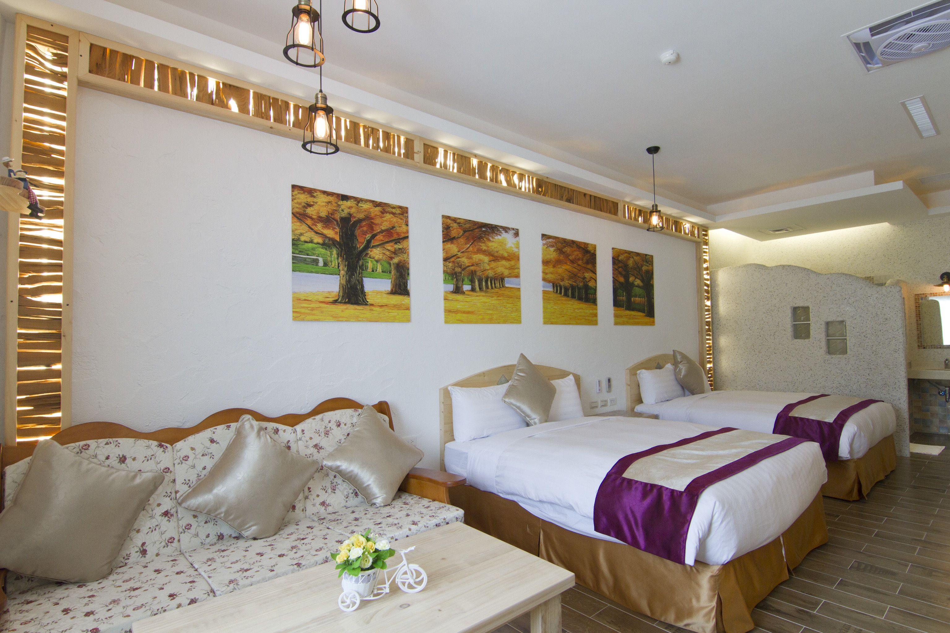 A hotel room with two beds, a sofa, a small table with a vase and a bicycle, and a wall with three autumn-themed paintings.