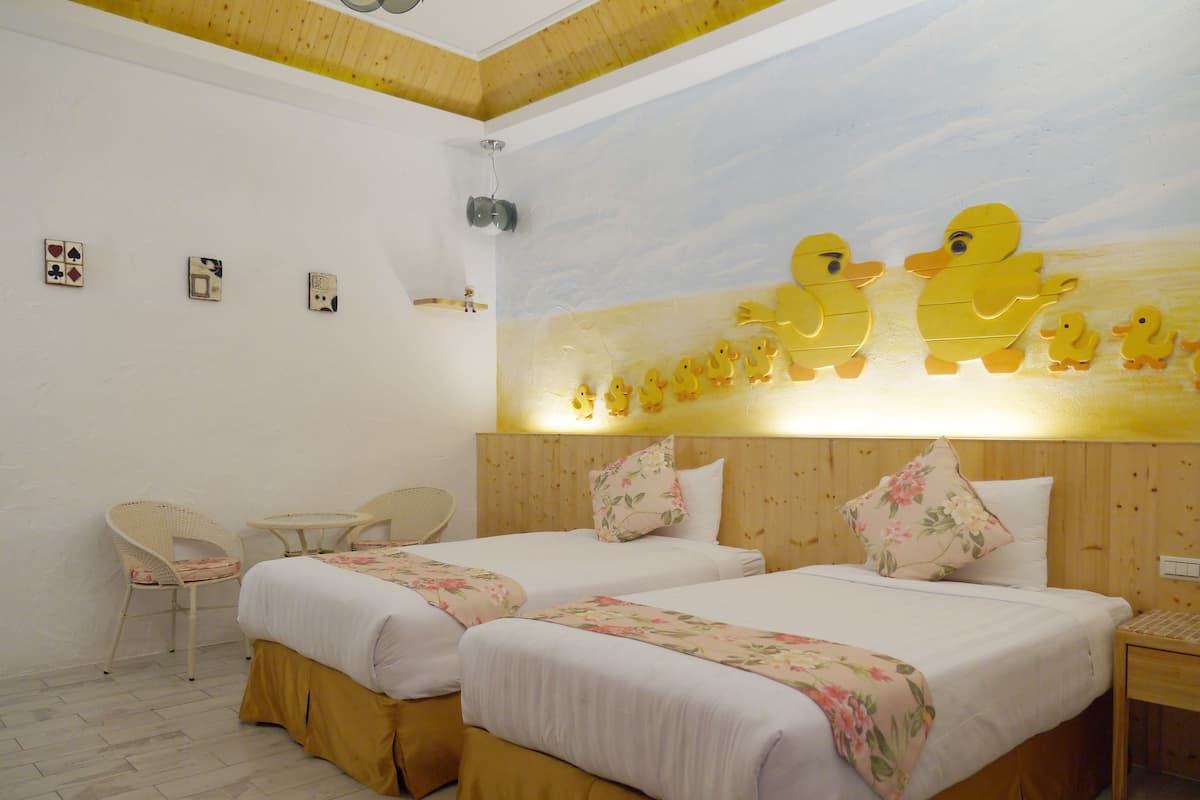 A hotel room with two beds, a wooden ceiling, a wall mural of yellow ducks, and a small table with chairs.