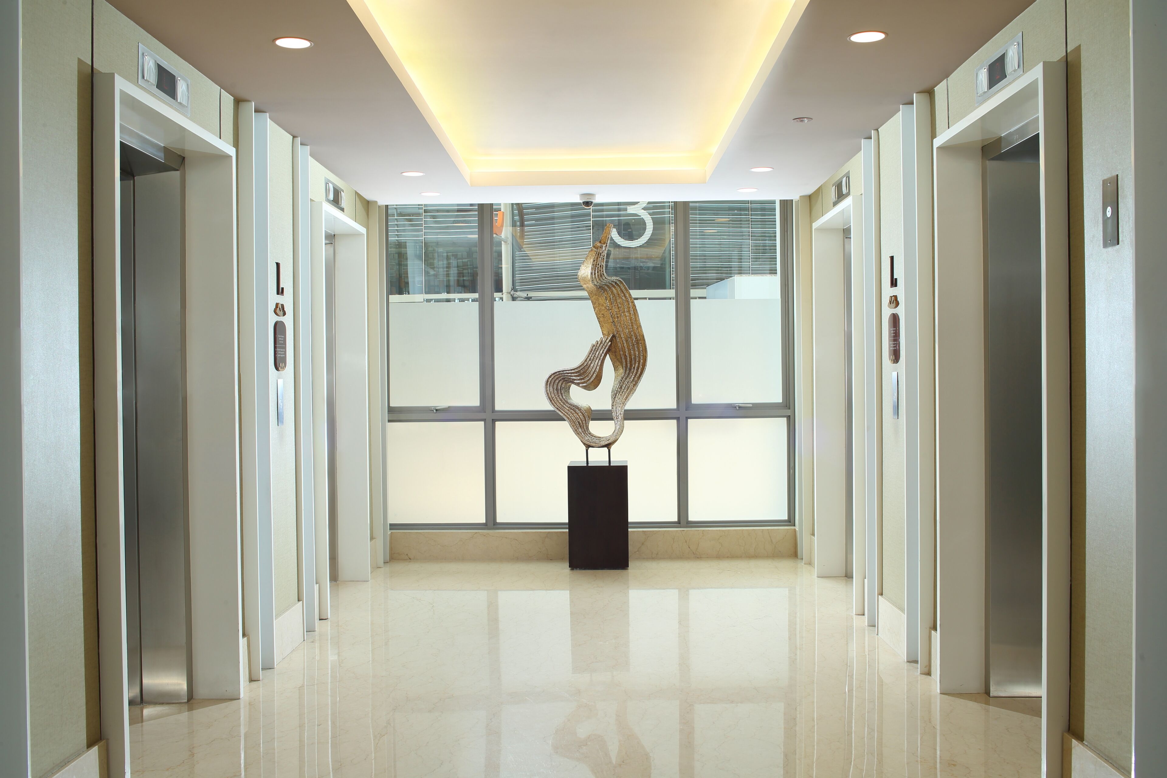Un moderno vestíbulo de ascensores con una escultura central, paredes de vidrio y piso pulido.