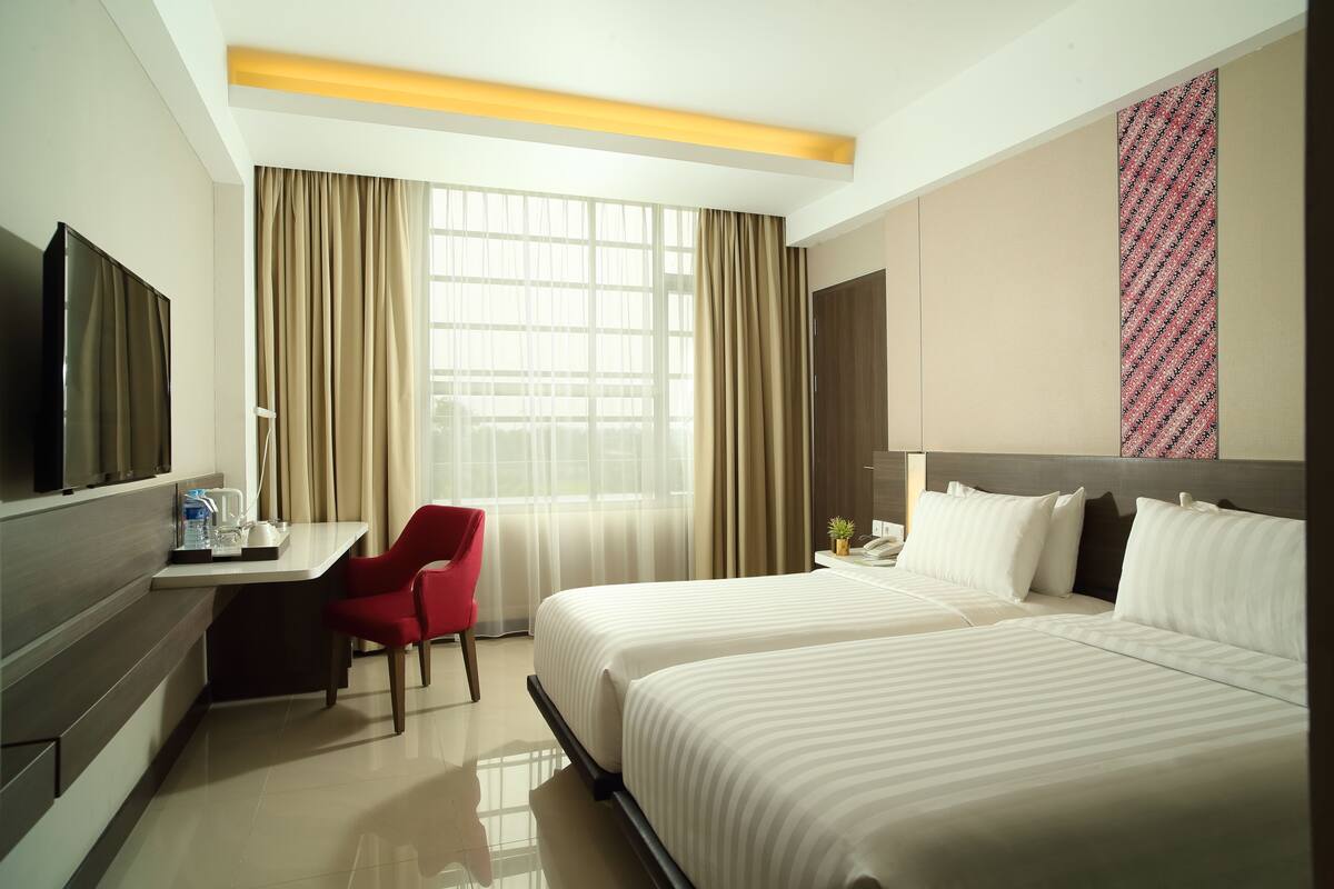 A hotel room with two beds, a desk, a TV, and a large window with curtains.