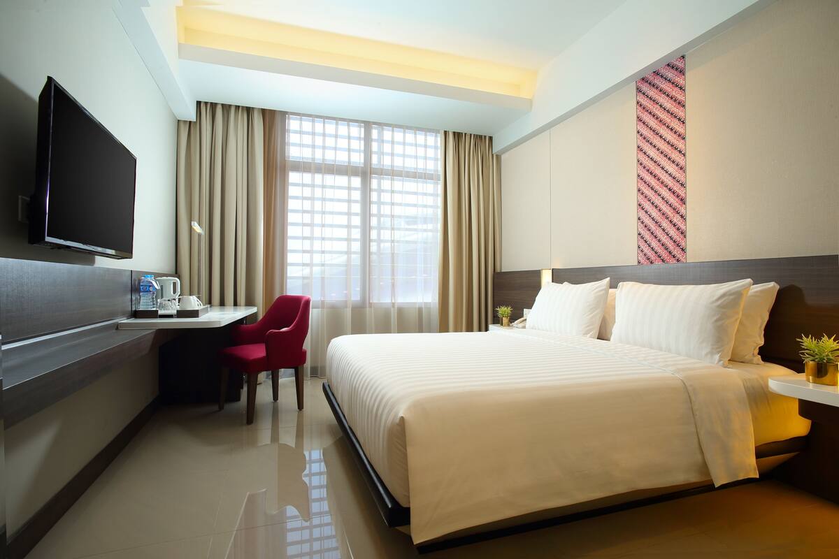 A hotel room with a large bed, a desk with a chair, a flat-screen TV, and a window with curtains.