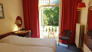 A hotel room with a bed, a desk with a lamp, a chair, and a view of the outdoors through a window with red curtains. - Nol in 't Bosch (Wageningen)