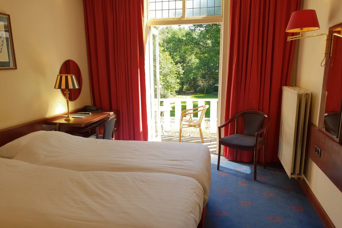A hotel room with a bed, a desk with a lamp, a chair, and a view of the outdoors through a window with red curtains.