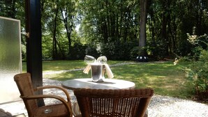 An outdoor patio with wicker furniture and a table set for two, surrounded by a lush garden and tall trees. - Nol in 't Bosch (Wageningen)