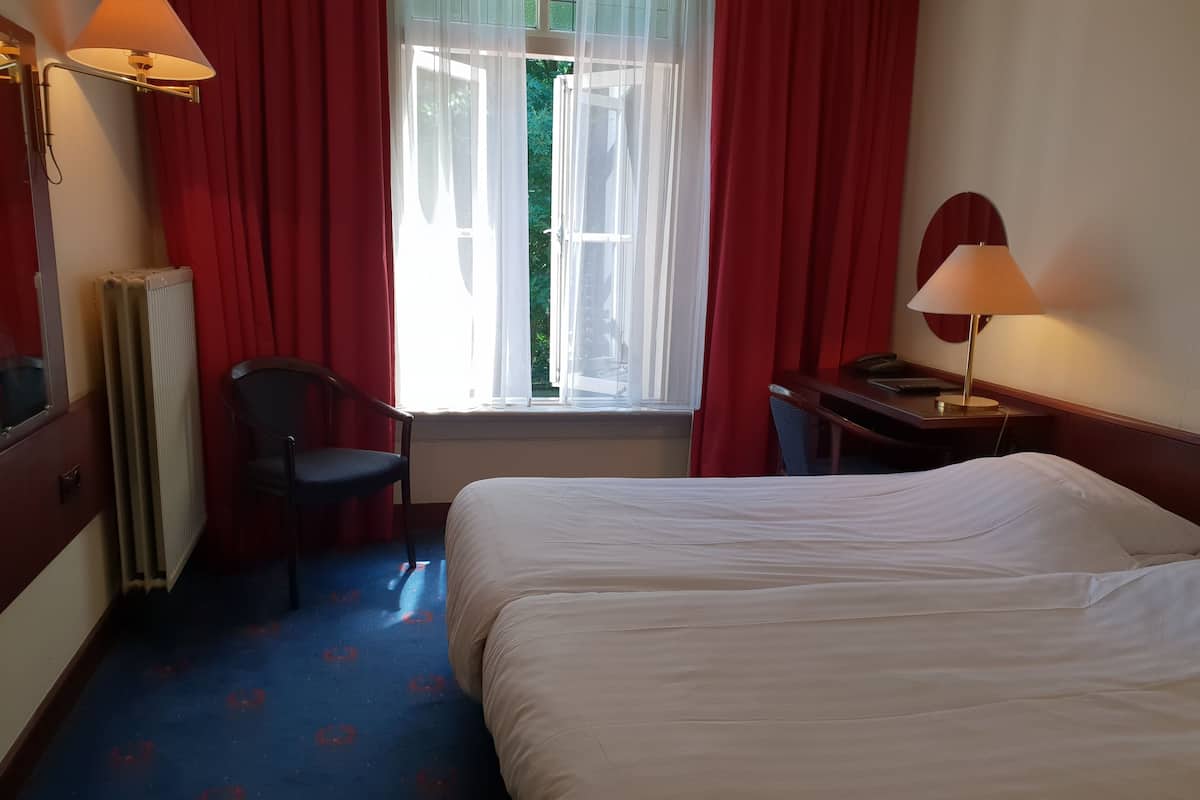 A hotel room with a bed, a desk, a chair, a radiator, and a window with curtains.
