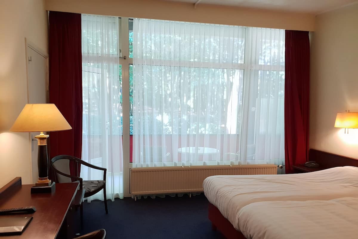 A hotel room with a bed, a desk with a lamp, a chair, a window with curtains, and a view of trees outside.