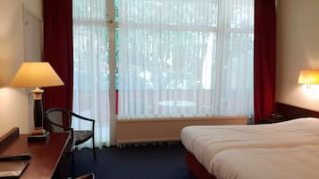 A hotel room with a bed, a desk with a lamp, a chair, a window with curtains, and a view of trees outside.