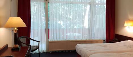 A hotel room with a bed, a desk with a lamp, a chair, a window with curtains, and a view of trees outside.