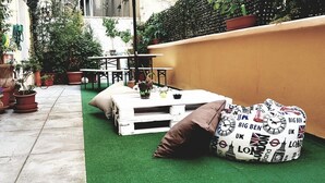 A patio area with a white table, chairs, and a green artificial turf. There are potted plants and a white wall in the background. - Smart Rooms (Trieste)