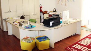 A reception desk with a computer, printer, and a fire extinguisher. There are two colorful stools and a coffee machine on the desk. - Smart Rooms (Trieste)