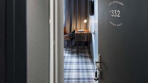 A hotel room with a checkered floor, a bed, a desk with a lamp, and a TV on the wall. - Hôtel Le Beaulieu (Charbonnieres-les-Bains)