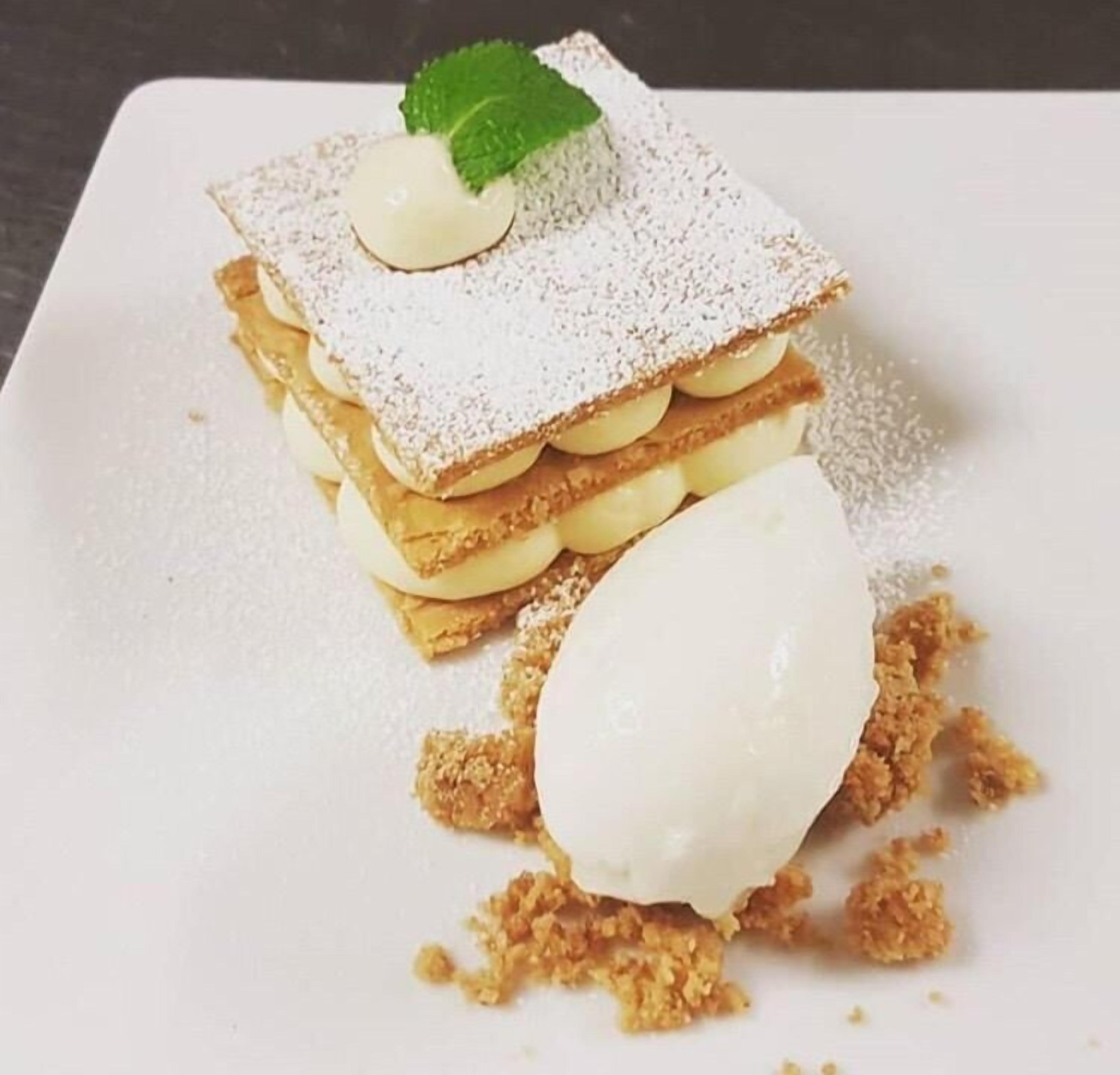 Un postre amb capes de pastí i crema, daurat amb sucre glas, i una fulla de menta com a guarnició.