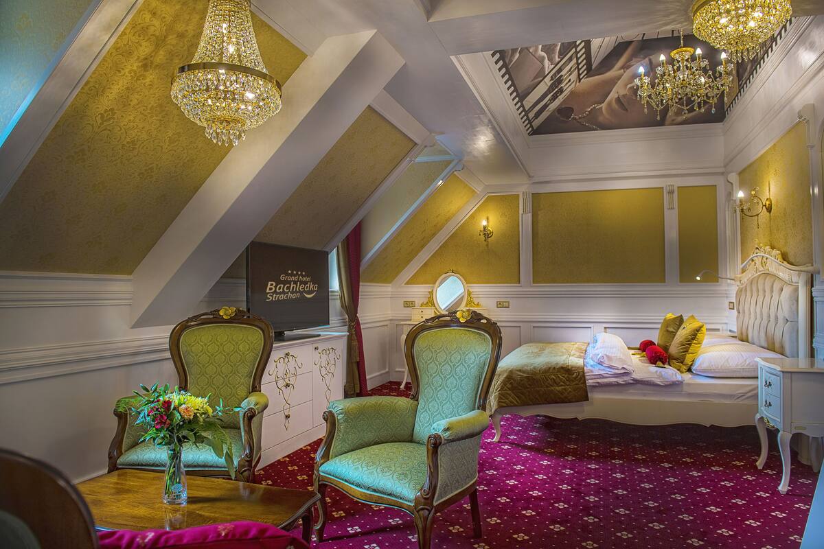 Une chambre à coucher luxueuse, dotée d’un grand lit, de fauteuils richement décorés, d’un lustre et d’une fresque peinte au plafond.