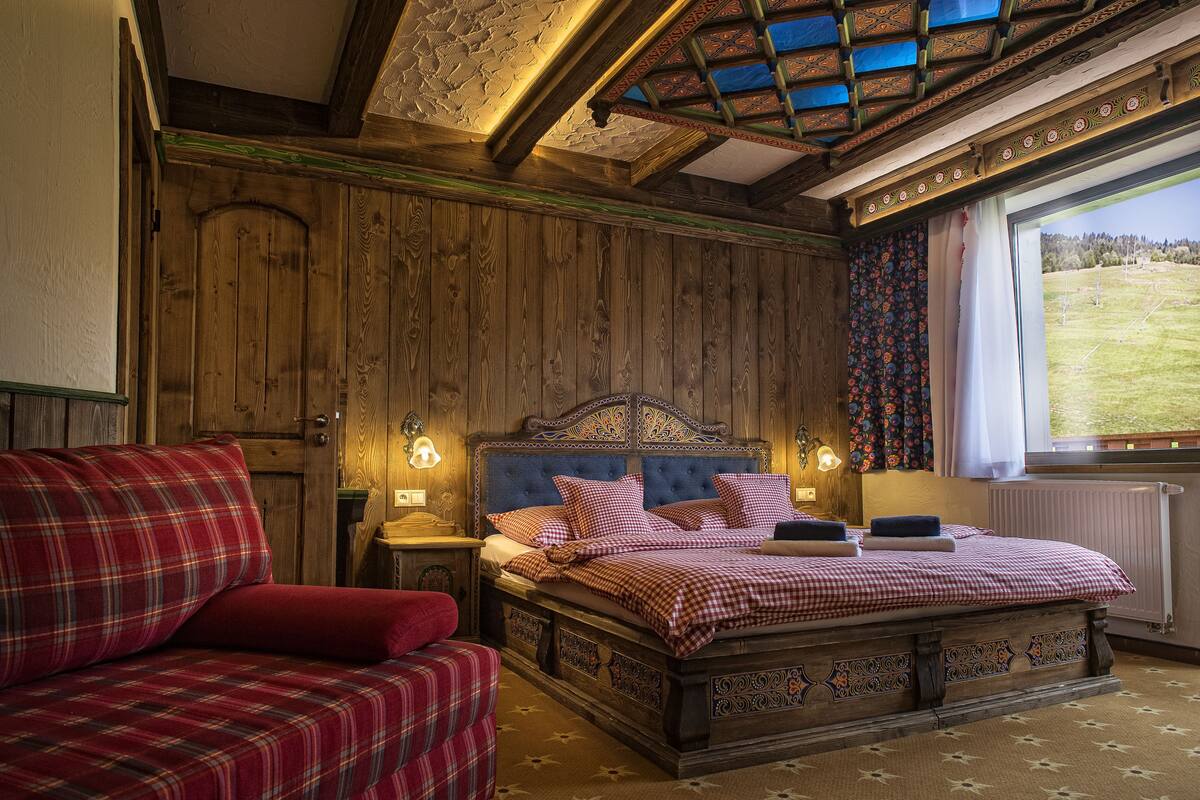 Une pièce traditionnelle en bois, avec un lit, un canapé à carreaux rouges et une fenêtre donnant sur un paysage verdoyant.