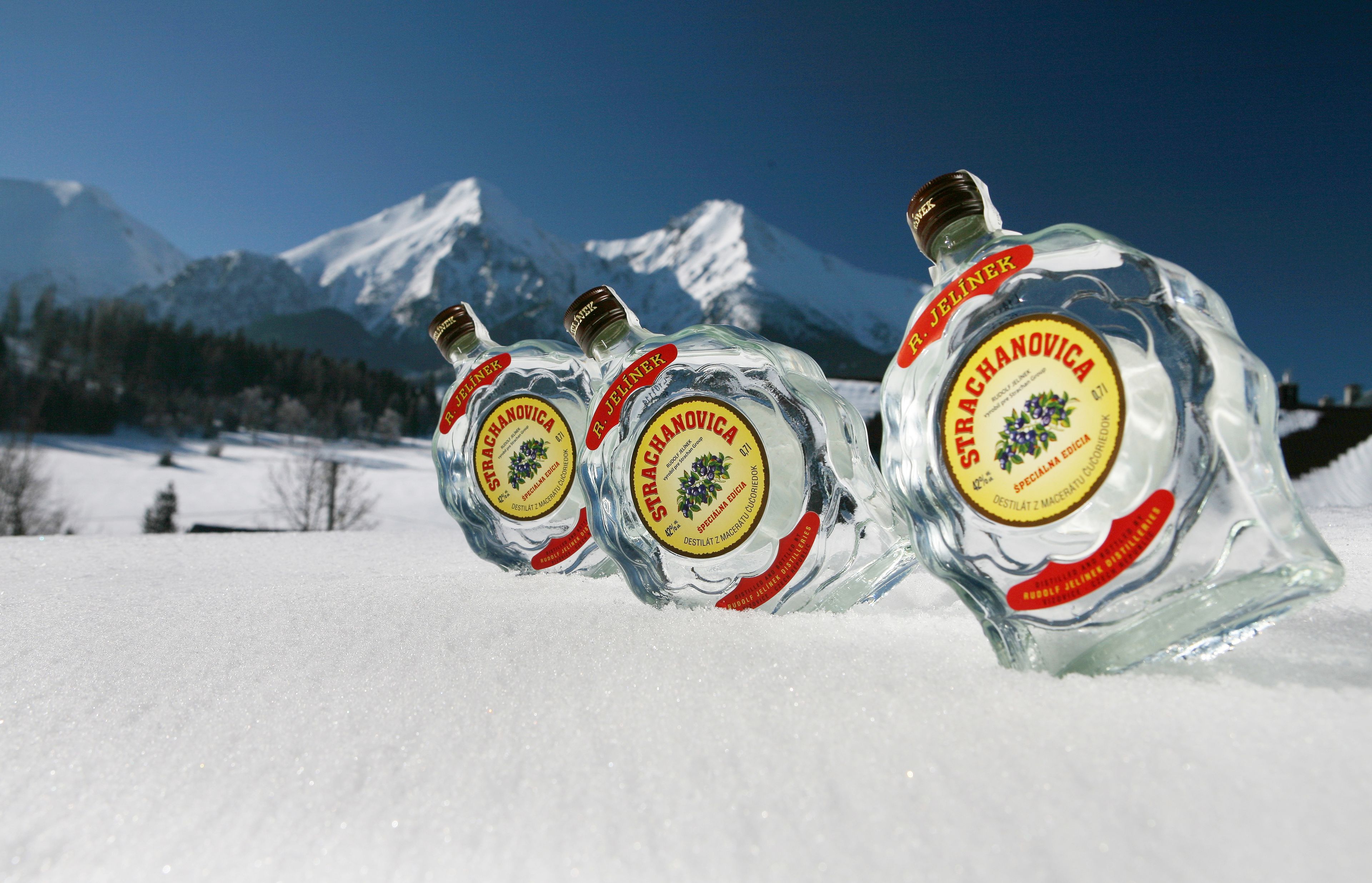 Tres botellas de vodka Strachanovica sobre una superficie nevada, con montañas al fondo.