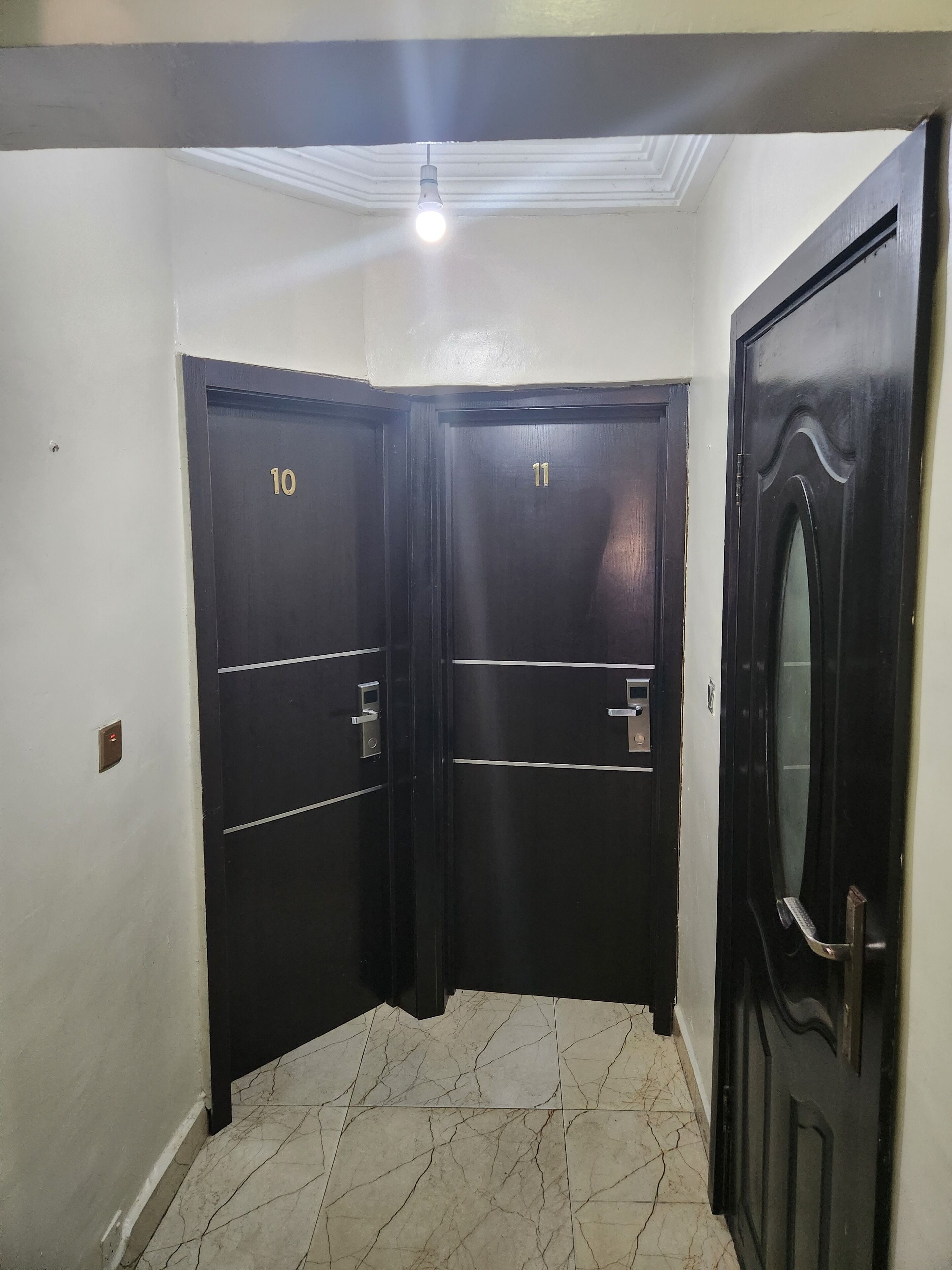 Un coin de couloir avec deux portes en bois sombre, numérotées 10 et 11, un luminaire au-dessus et un sol carrelé.
