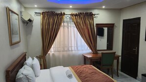 Quarto de hotel com cama, escrivaninha, cadeira, televisão e porta.