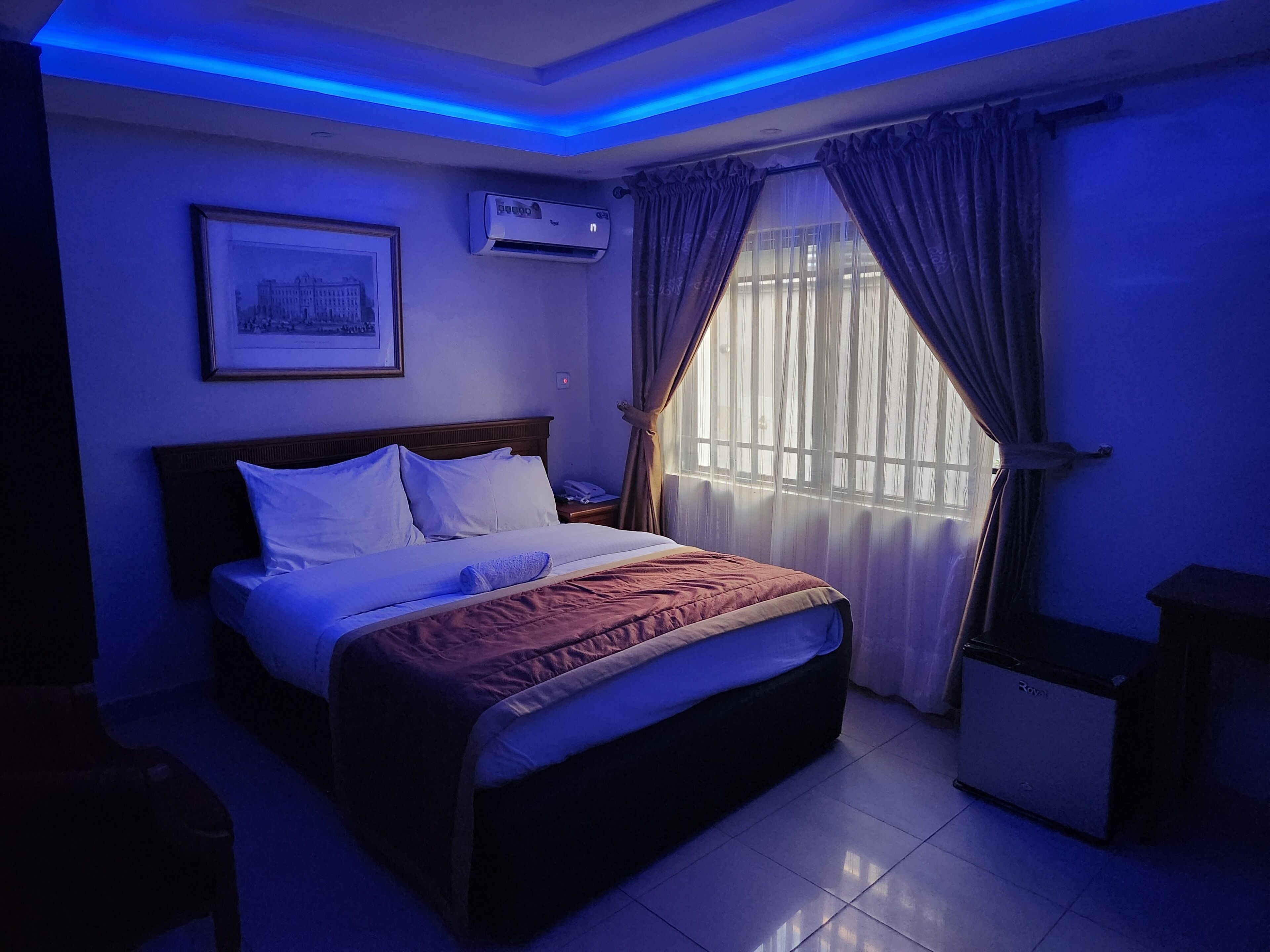 A hotel room with a bed, an air conditioning unit, a framed picture on the wall, and a window with curtains.