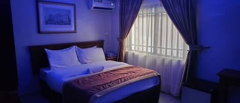 Une chambre d’hôtel avec un lit, un appareil de climatisation, un tableau encadré au mur et une fenêtre avec des rideaux.