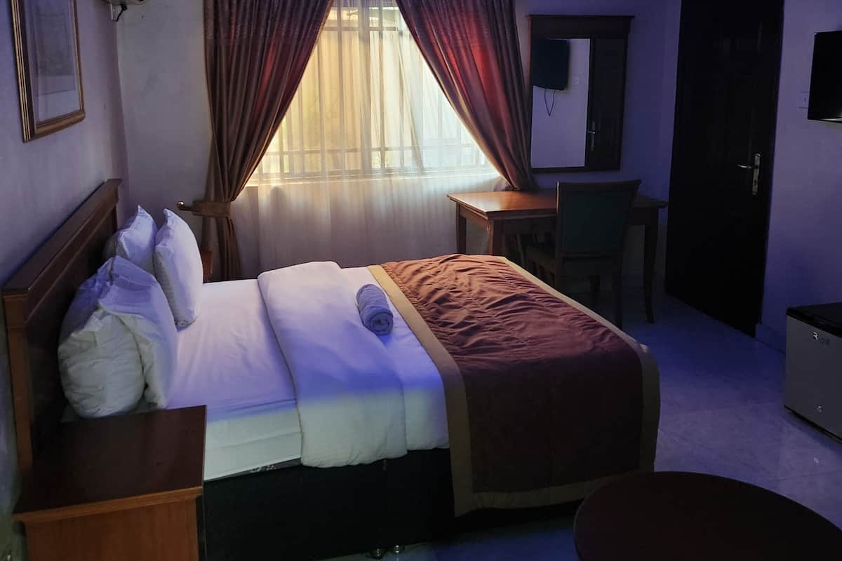 A hotel room with a bed, bedside table, chair, desk, and a TV on the wall.