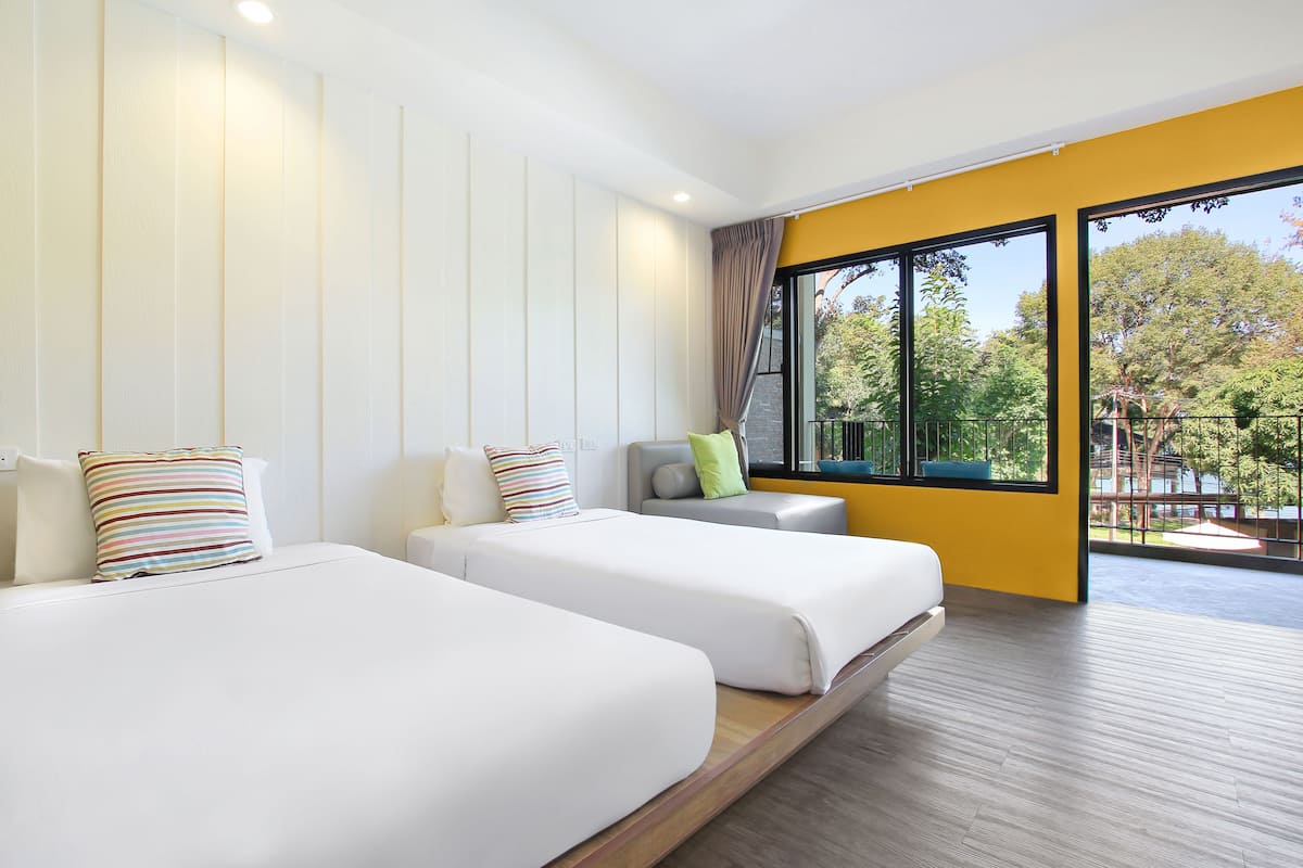 Quarto de hotel moderno com duas camas, uma janela grande com vista para vegetação e uma parede de destaque amarela.