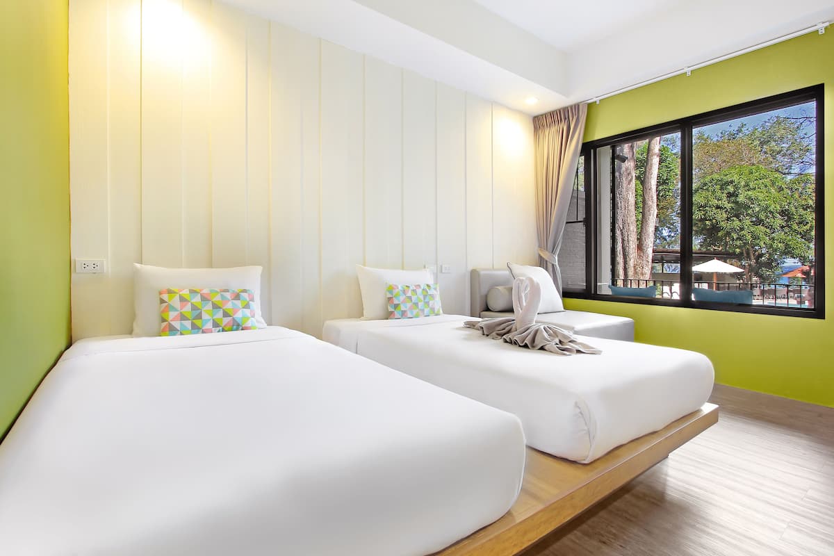 Quarto de hotel com duas camas, uma janela com vista para árvores e guarda-sóis, e piso de madeira.