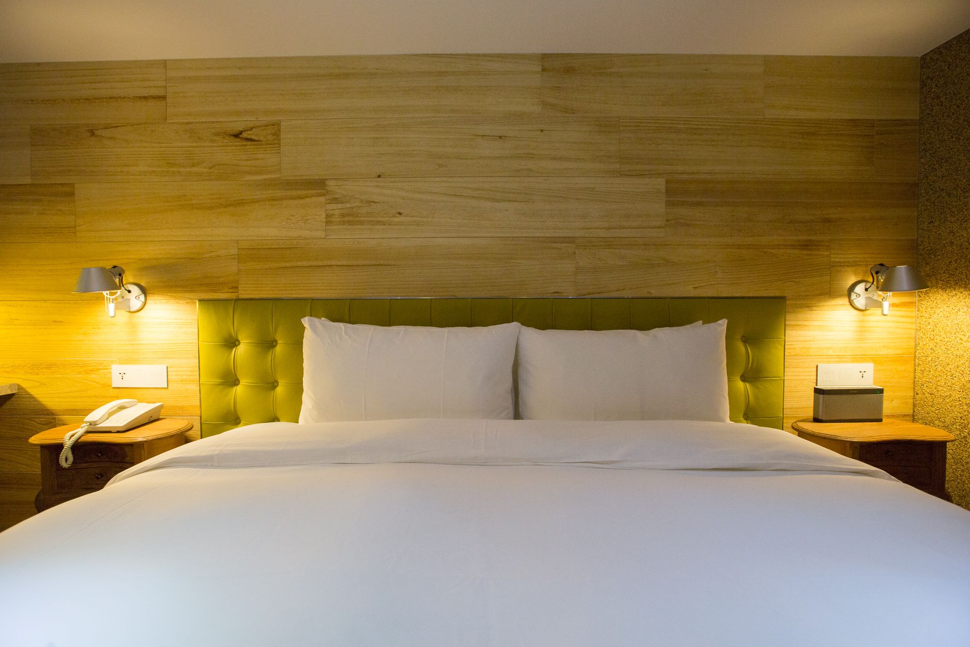 Une chambre d’hôtel avec une tête de lit recouverte d’un tissu vert, une couvre-lit blanche, deux lampes de chevet et un téléphone sur la table de chevet.