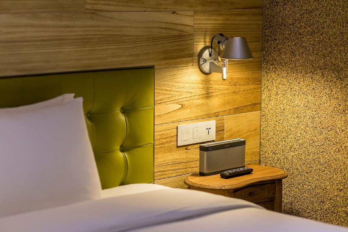 A hotel room with a green upholstered headboard, a wooden wall panel, a wall-mounted lamp, a power outlet, a small wooden nightstand, and a remote control.