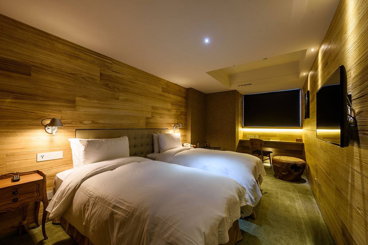 A hotel room with two beds, wooden paneling on the walls, a flat-screen TV mounted on the wall, and a small table with a chair.