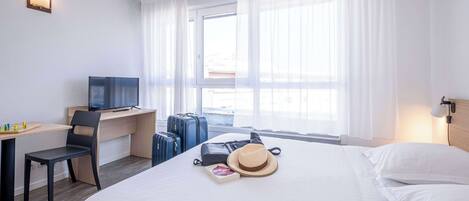 Habitación de hotel con cama, escritorio con televisor, una silla y ventana con cortinas.