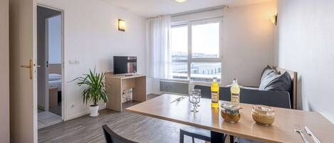 Habitación de hotel moderna con zona de comedor, televisor, planta en maceta y ventana con vista a la ciudad.