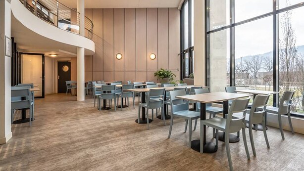 A modern dining area with multiple tables and chairs, large windows offering a view of the outdoors, and a curved staircase leading to an upper level. - Comfort Aparthotel Les Hauts d'Annecy (Annecy)