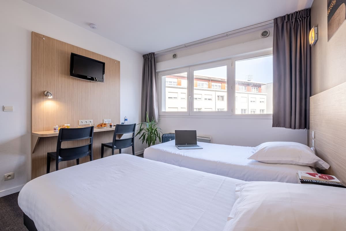 Habitación de hotel con dos camas, un escritorio con una computadora portátil, un televisor en la pared y una ventana con cortinas.