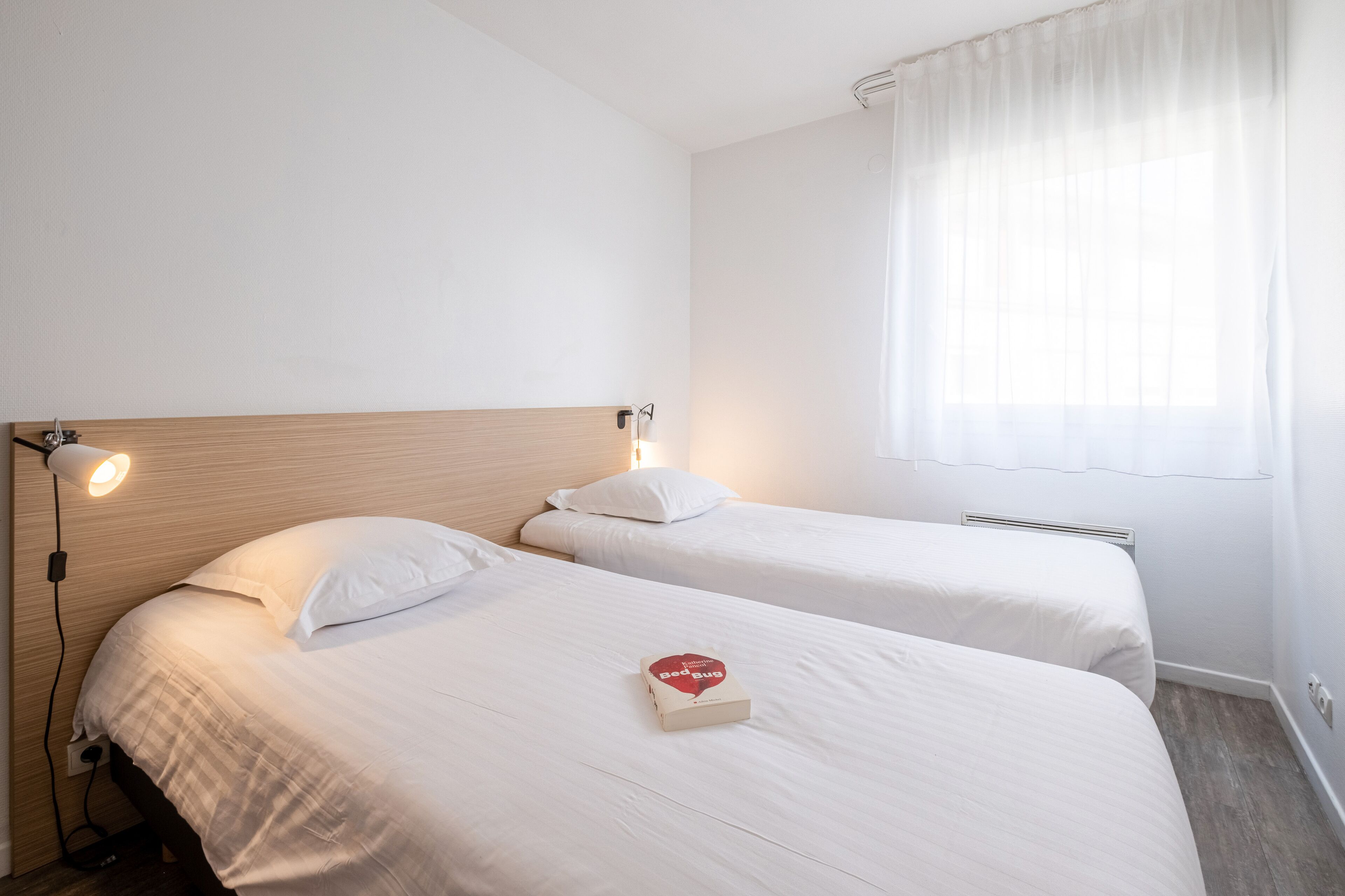 Habitación de hotel con dos camas individuales, una mesita de noche con lámpara y una ventana con cortinas traslúcidas.