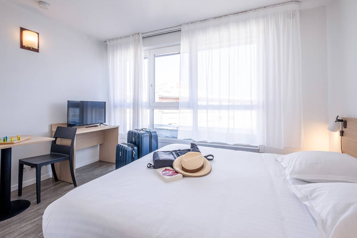 Habitación de hotel con cama, escritorio con computadora, silla, televisor y ventana con cortinas.