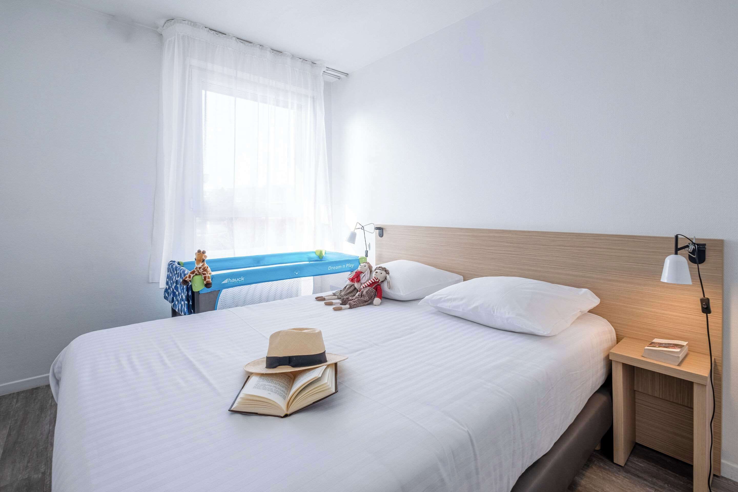 A hotel room with a bed, a nightstand, a lamp, a window with curtains, and a view of a blue sky.