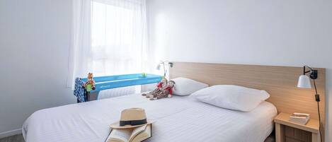 A hotel room with a bed, a nightstand, a lamp, a window with curtains, and a view of a blue sky.