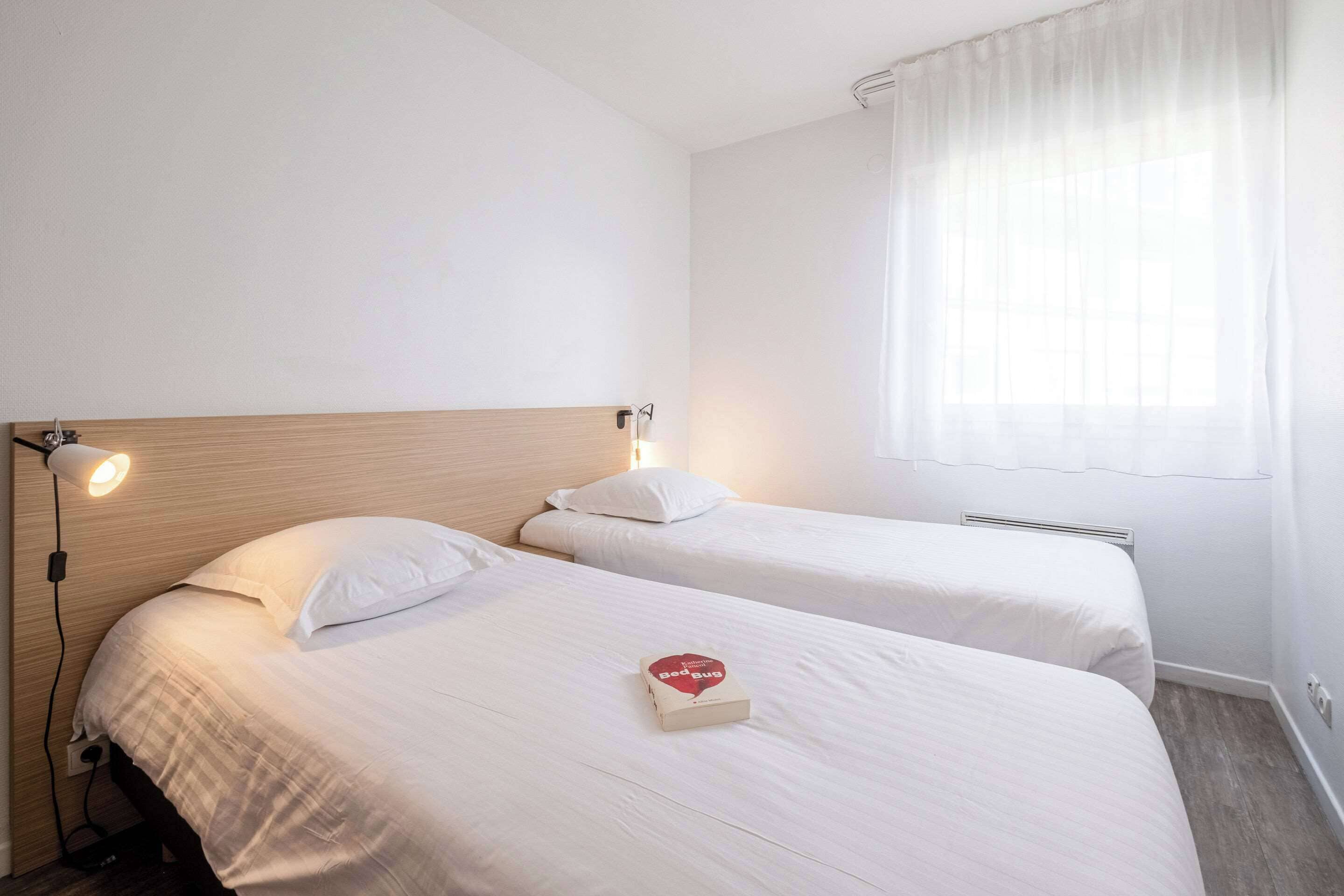 A hotel room with two beds, a bedside lamp, a window with sheer curtains, and a bedside table with a book.