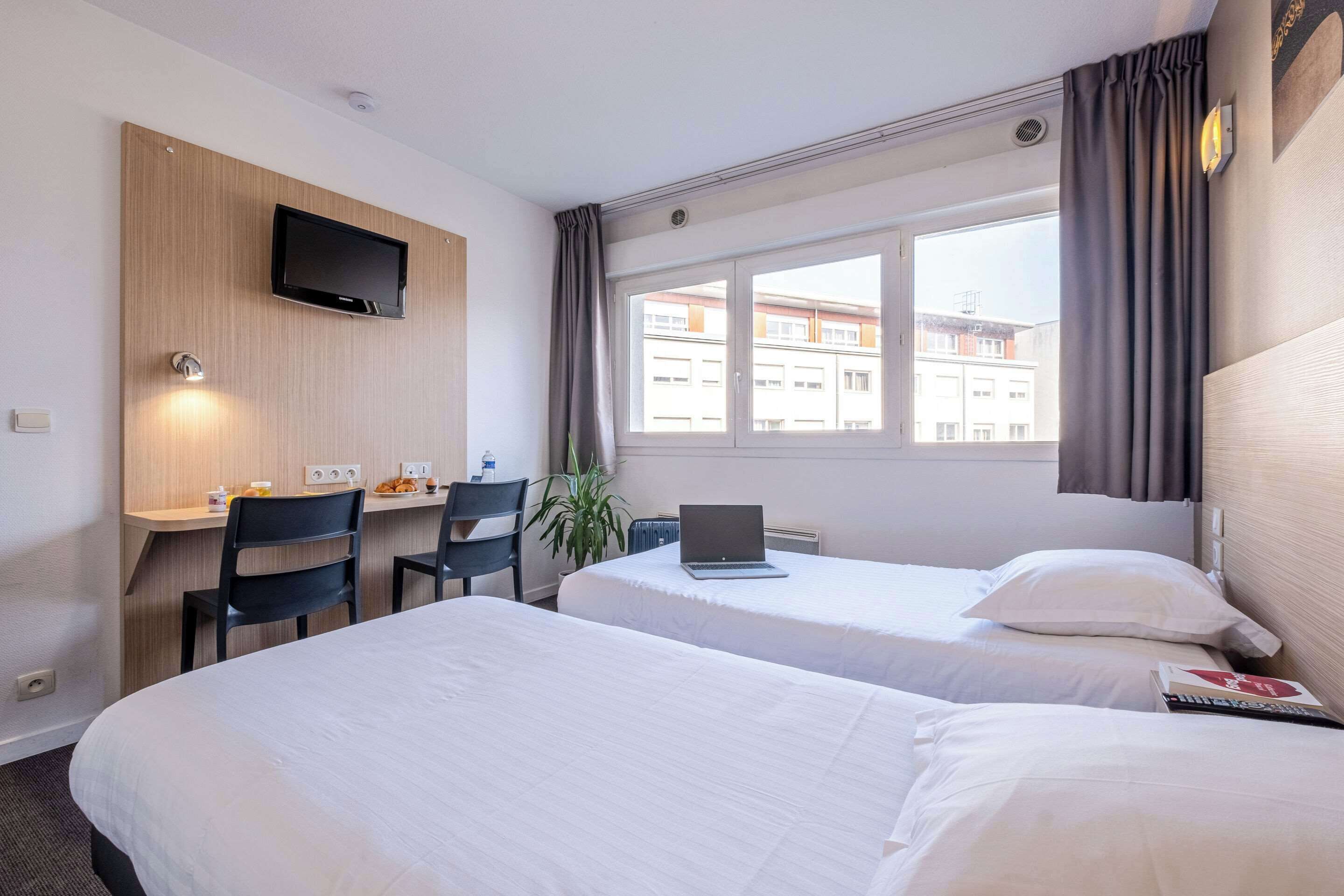 A hotel room with two beds, a desk with a laptop, a TV mounted on the wall, and a window with curtains.