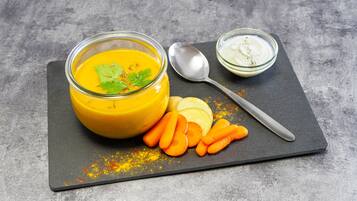 A bowl of yellow soup with a garnish, a spoon, and a small bowl of white sauce on a slate board with carrot sticks.