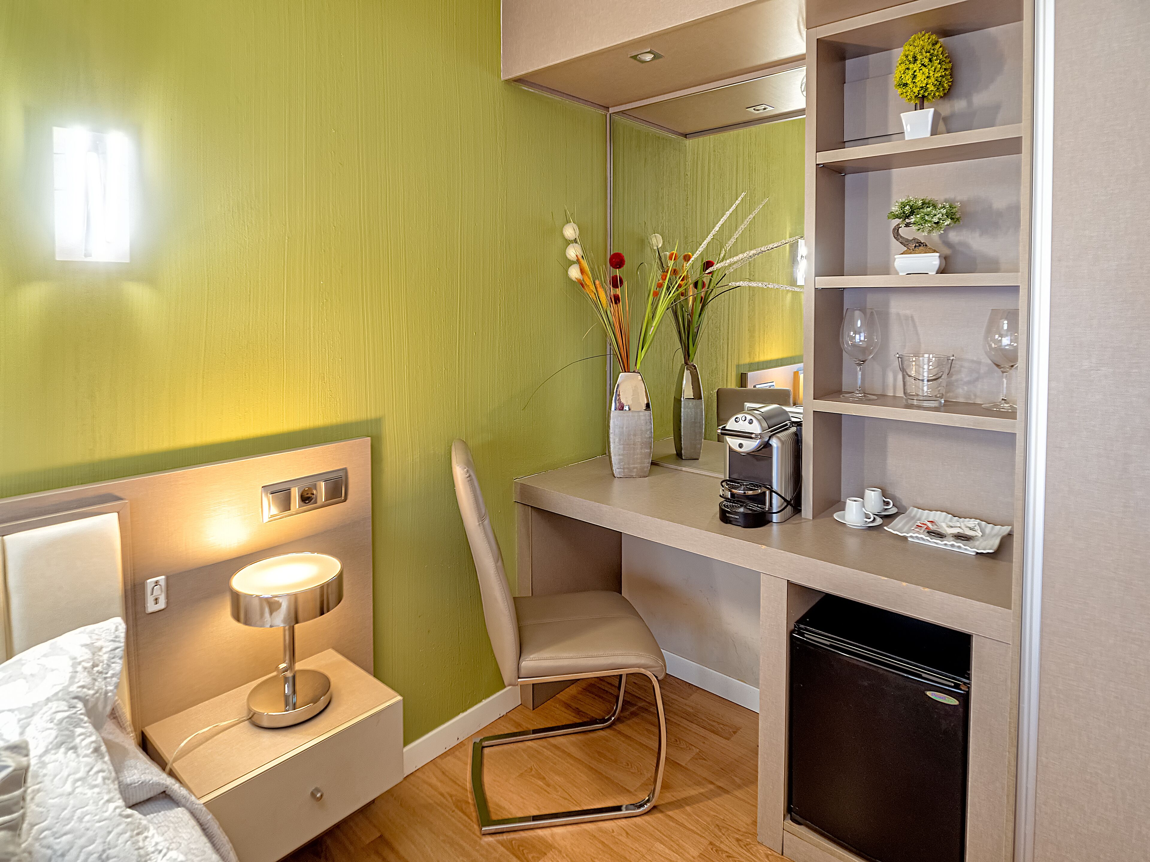 Ein gemütliches Hotelzimmer mit Bett, Schreibtisch mit Kaffeemaschine, Stuhl, kleinem Kühlschrank und Ablagefläche mit Dekoration.