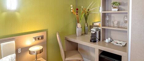 A compact hotel room with a bed, a desk with a coffee maker, a chair, a small refrigerator, and a shelf with decorative items.