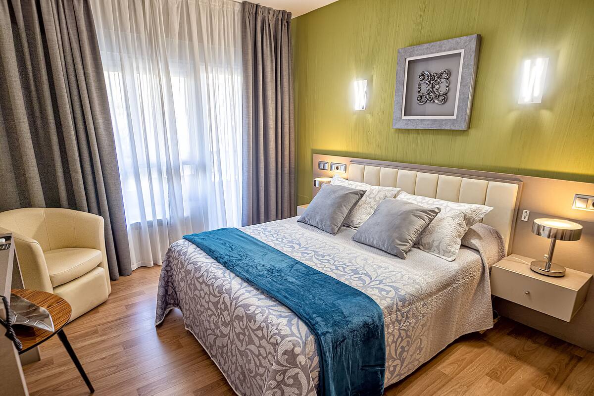 A neatly arranged bedroom with a bed, a chair, a nightstand, and a wall-mounted light fixture.