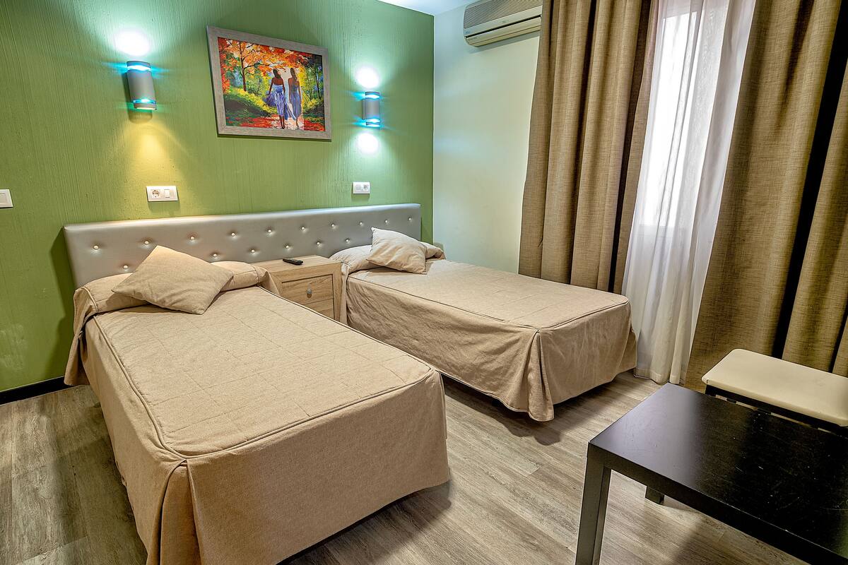A hotel room with two beds, a nightstand, a table, and a painting on the wall.