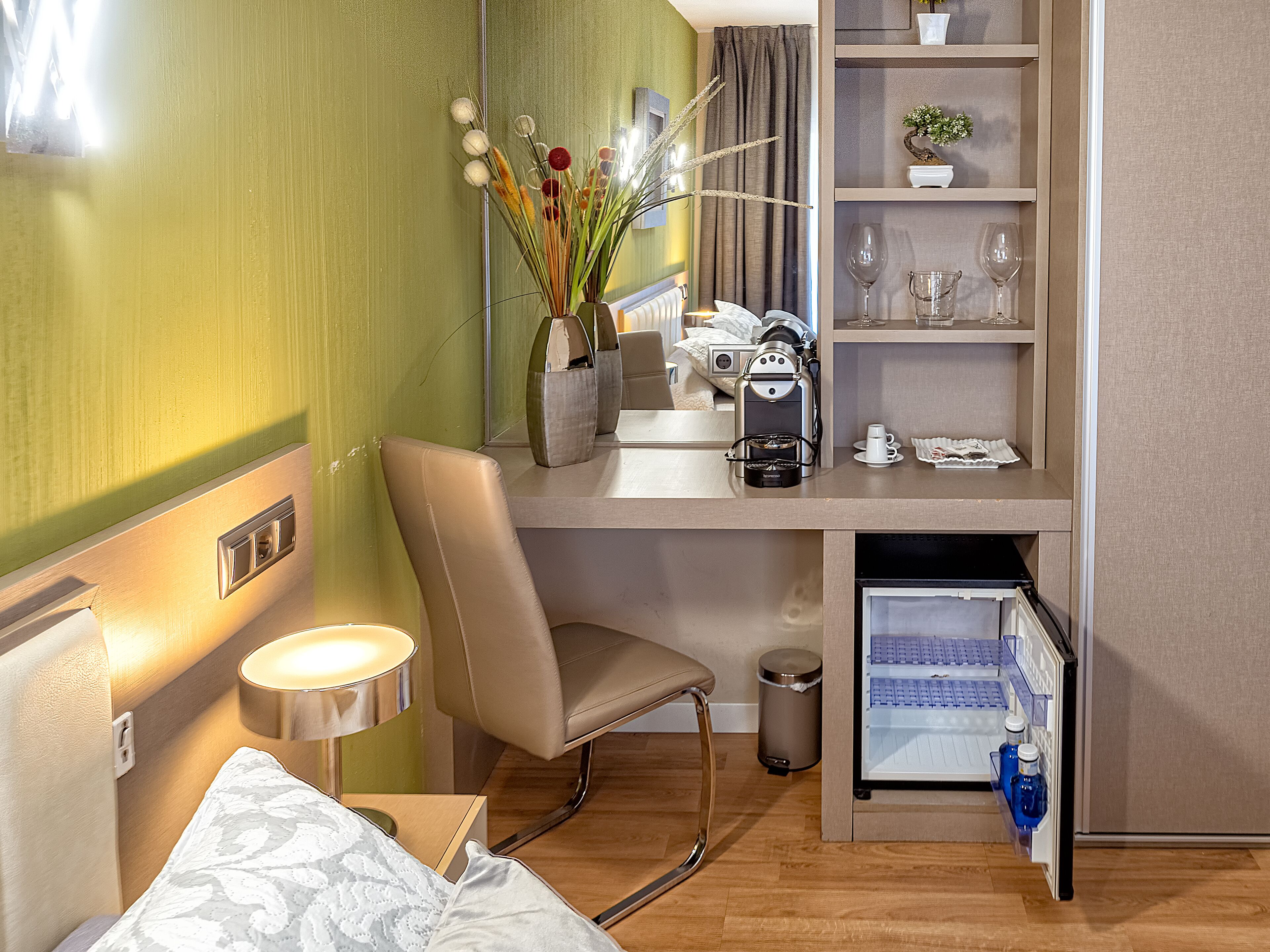 Ein kleines Hotelzimmer mit Kochnische, einem Bett mit gemustertem Kopfkissen, einem Stuhl, einem kleinen Mülleimer und einem Minikühlschrank mit Getränken.