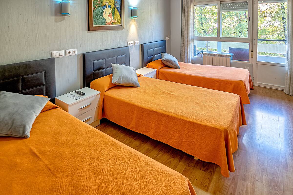 A hotel room with three single beds, a nightstand, a wall-mounted light, and a window with a view of trees.